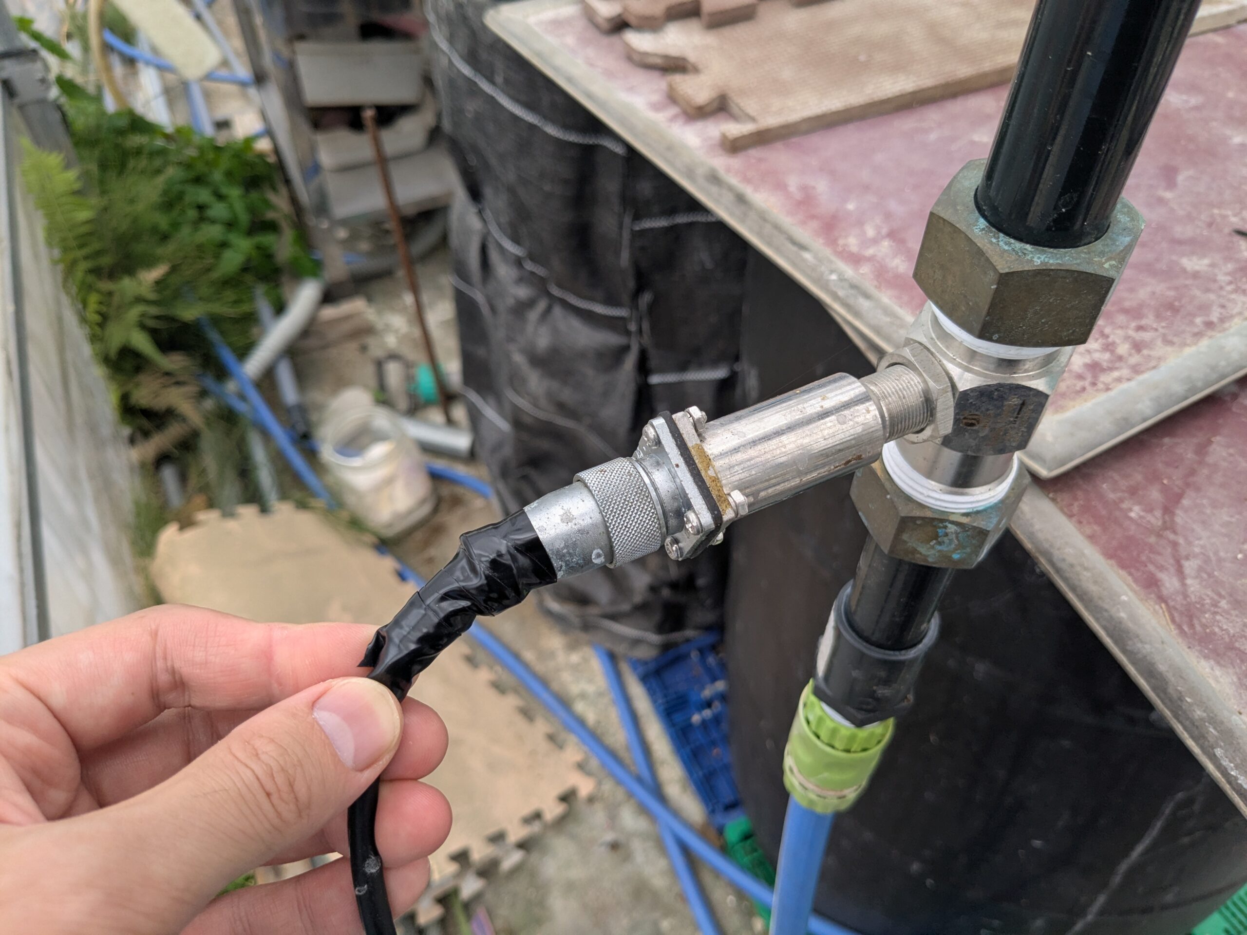 黒い配管のT字ユニオンに横付けされた銀色の流量センサー。手前にケーブル側のコネクタを持っている作業前の様子